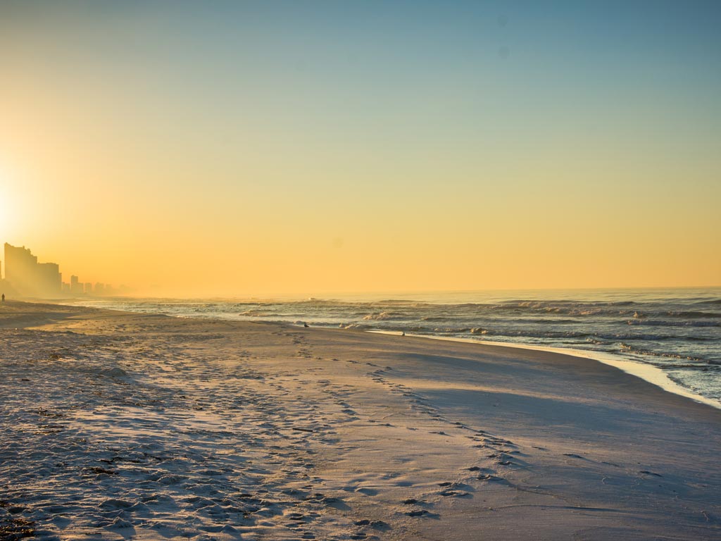 A sunset shot of a beach in Panama City Beach, Florida, with foot traces visible in the sand, and waves slowly crashing against it. 