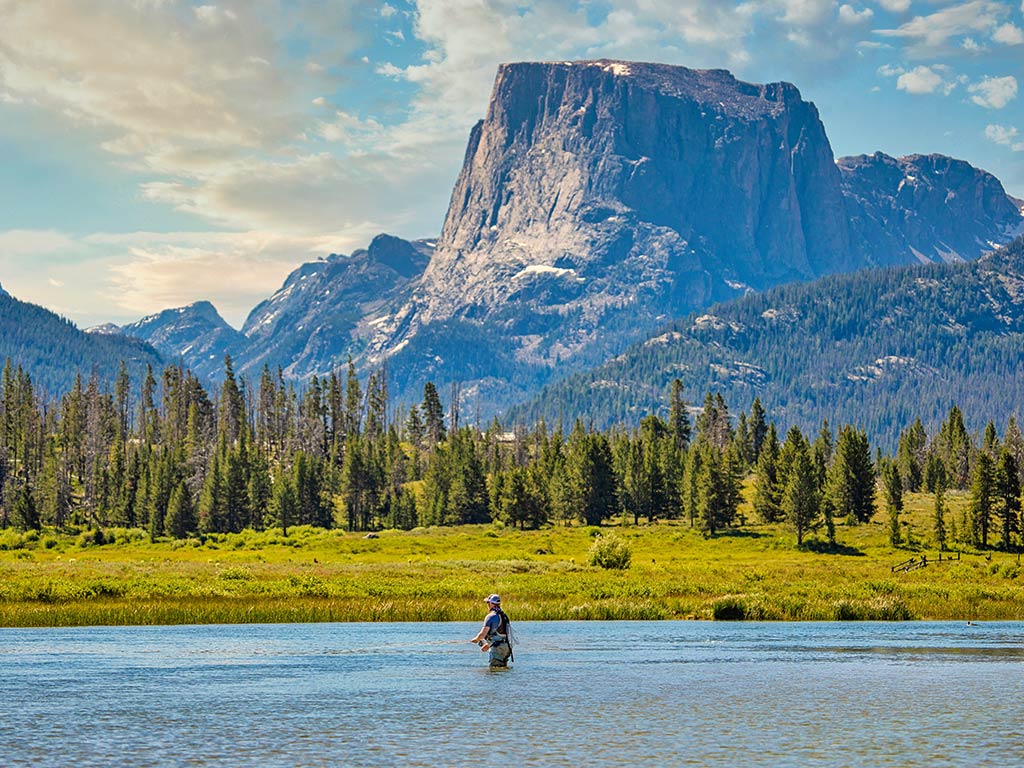 A view across a river towards a large rocky mountain in the distance on a clear day in Wyoming, with a sole fly angler casting their line in the water in the foreground