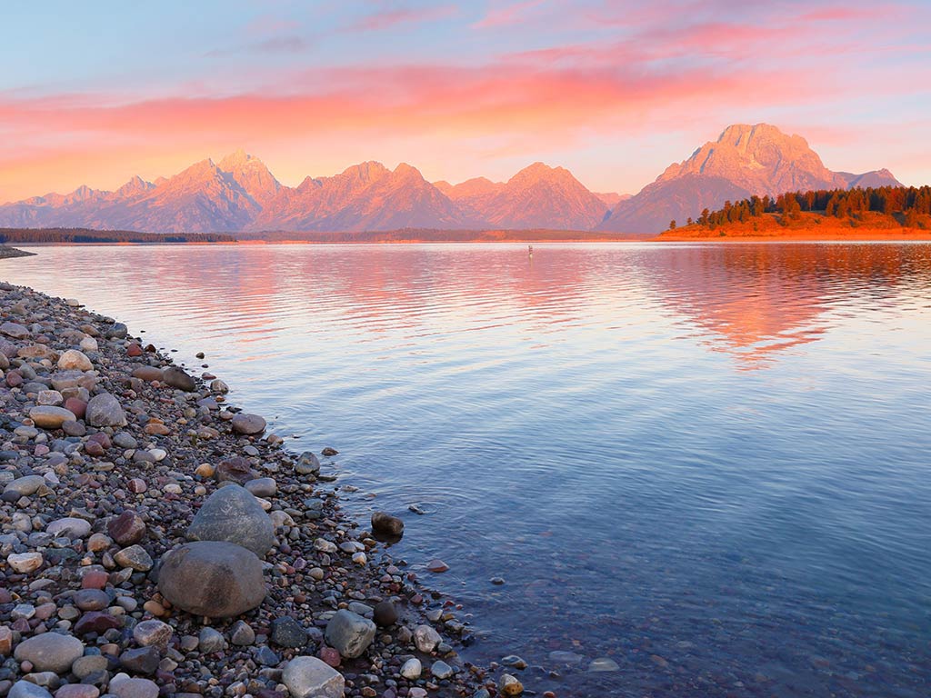 A view across the calm waters of Jackson Lake, Wyoming, at sunset with pink skies visible over some rocky mountains in the distance