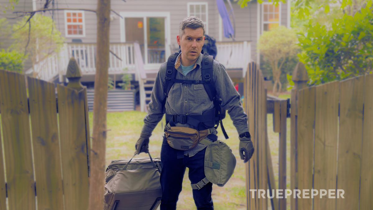 Sean pulling an INCH duffle through a gate while wearing a backpack, admin pouch, and gas mask carrier with his home behind him.
