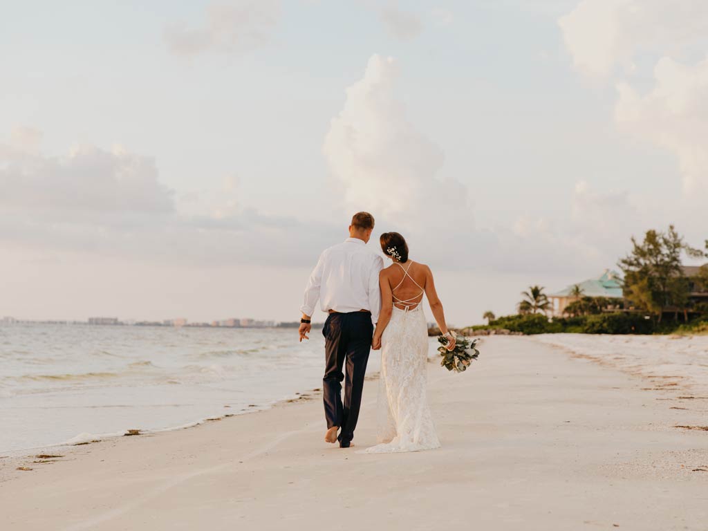 A photo featuring a couple who just got married walking peacefully while holding hands on a white sand beach during sunset