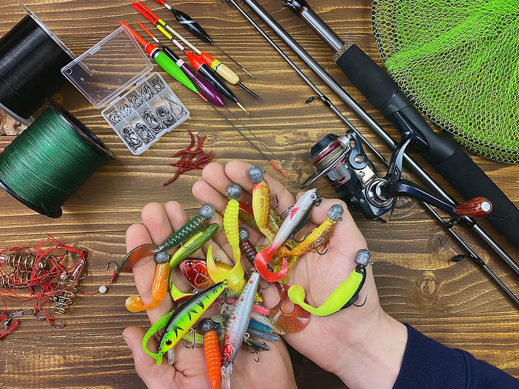 A view from above of two hands holding a selection of colorful jig fishing lures above a table full of fishing gear