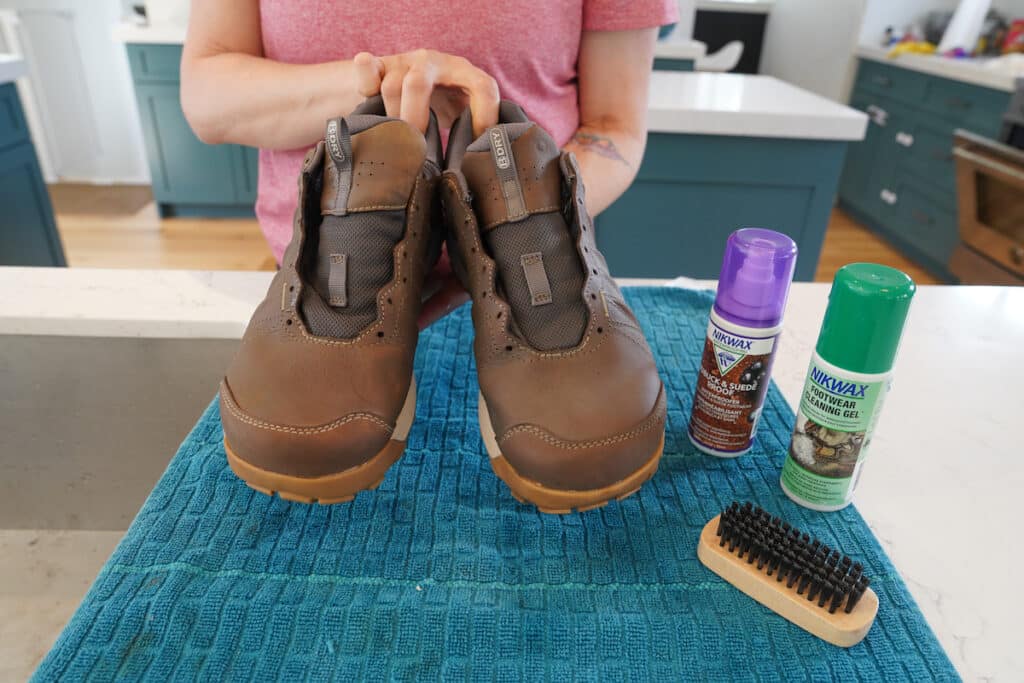 woman holding up a pair of cleaned wet hiking boots without the laces. Nikwax footwear cleaning gel and waterproofing spray are next to the shoes