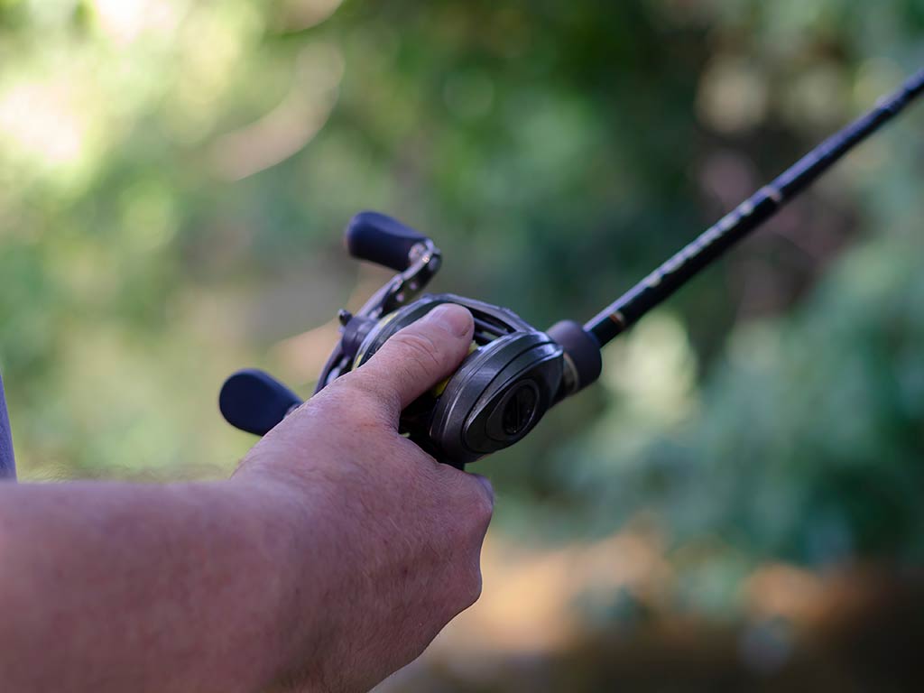 A closeup of a hand holding a baitcasting fishing rod and reel combo with the thumb partially covering the spool