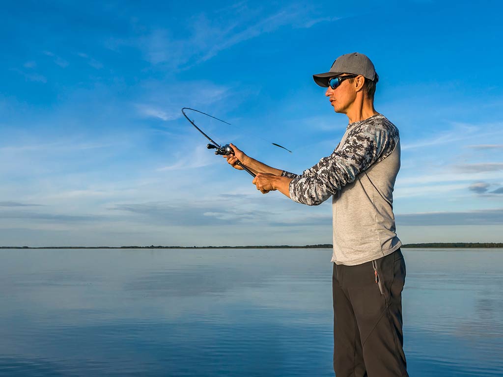 A man in a baseball cap and sunglasses casting a baitcasting rod and reel combo with a sideways action on a calm lake on a clear day