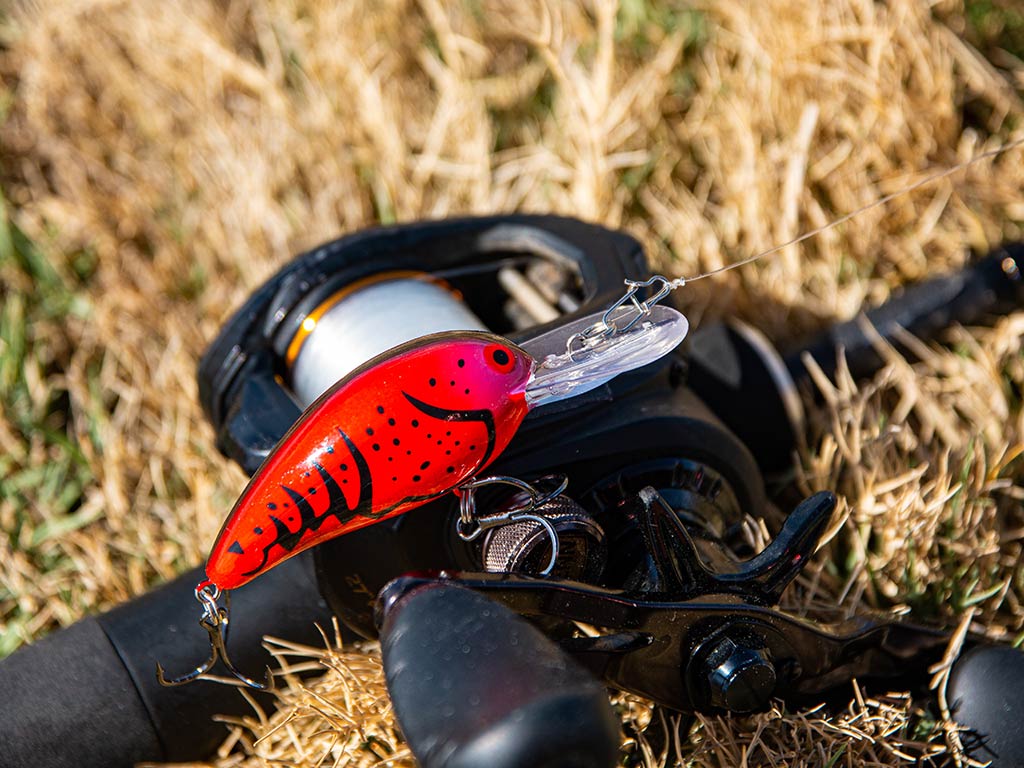 A closeup of a red fishing lure hanging next to a baitcasting fishing reel on the ground in some light-colored grass
