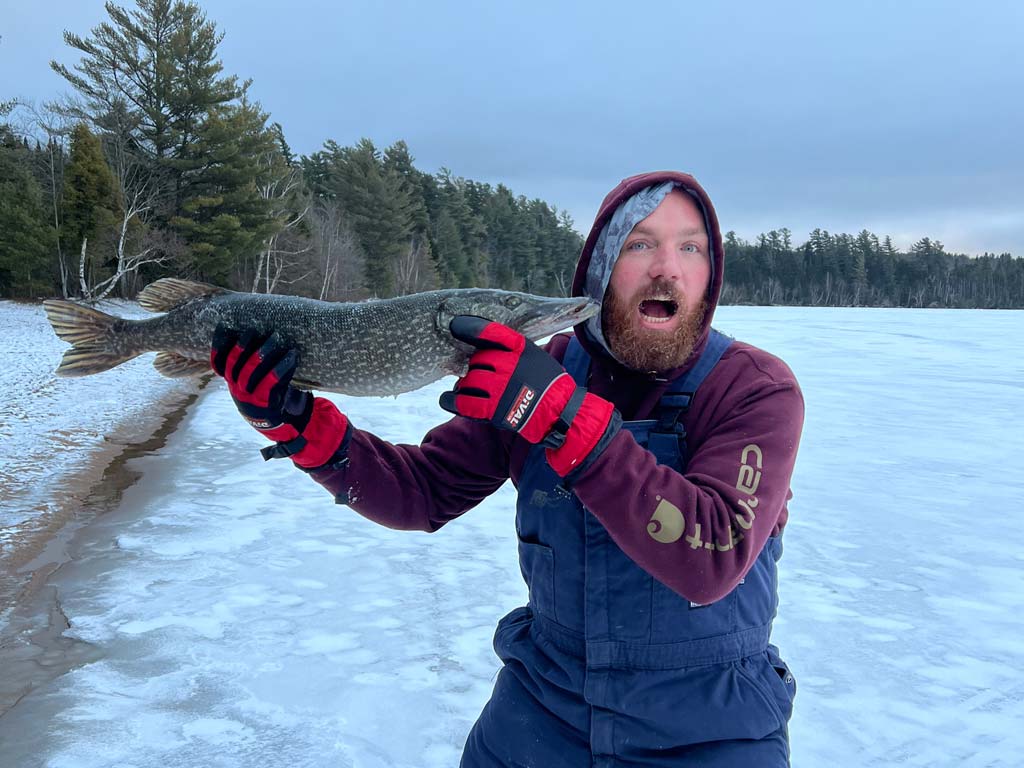 An angler making a scared face while holding a Northern Pike he caught ice fishing next to his head, with the snowed-over lake and coastal forests visible in the background.