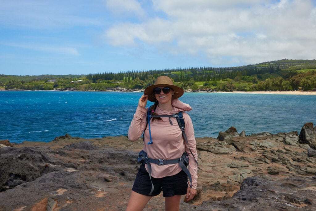 Woman hiking in front of the ocean in Maui wearing hiking shorts, REI Sahara Shade long sleeve shirt for sun protection, a hat, and a hiking daypack