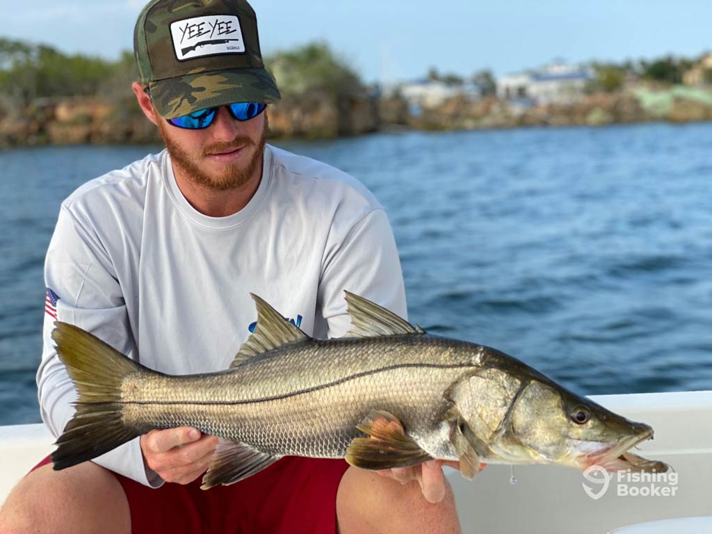 A photo featuring an angler wearing a cap and a pair of sunglasses while sitting on a charter boat and holding one of the most famous inshore fish species, Snook