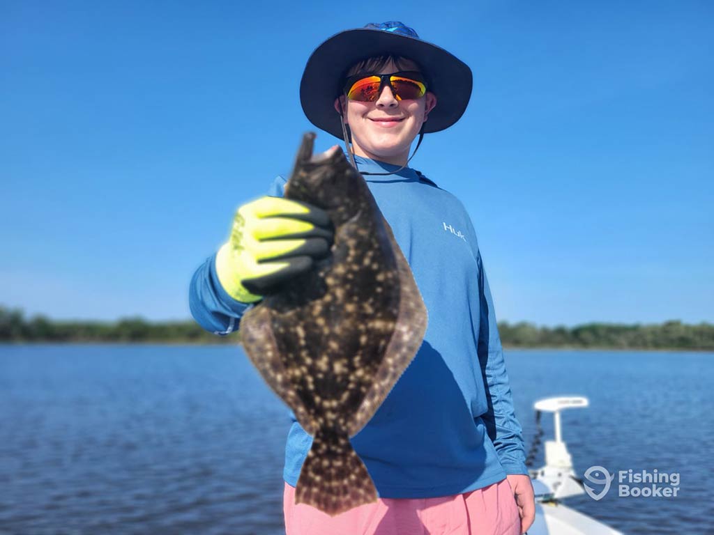 A child wearing a hat, sunglasses, and gloves holds up a Flounder to the camera aboard a fishing boat, with calm in shore waters behind him and blue skies above