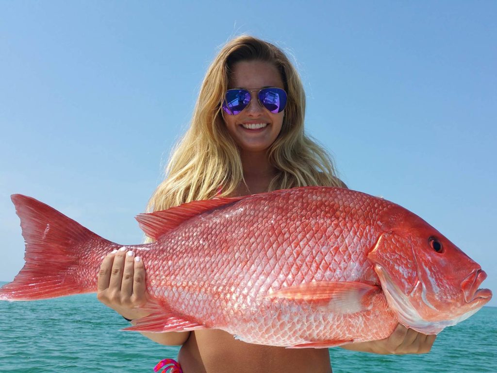 A smiling blonde female angler in sunglasses and a bathing suit, holding a large Red Snapper, with blue skies and water in the background