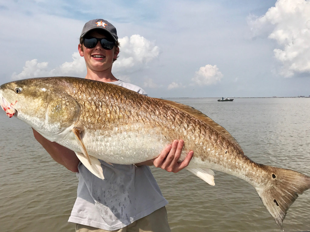 A happy and satisfied young angler struggling to hold a big Bull, almost state record-like Redfish with both hands while standing on a Texas charter fishing boat