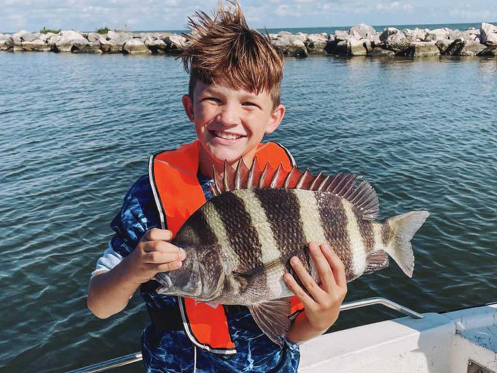 A photo of a young boy smiling while posing in front of the jetties with his Sheepshead on a beautiful and sunny spring day