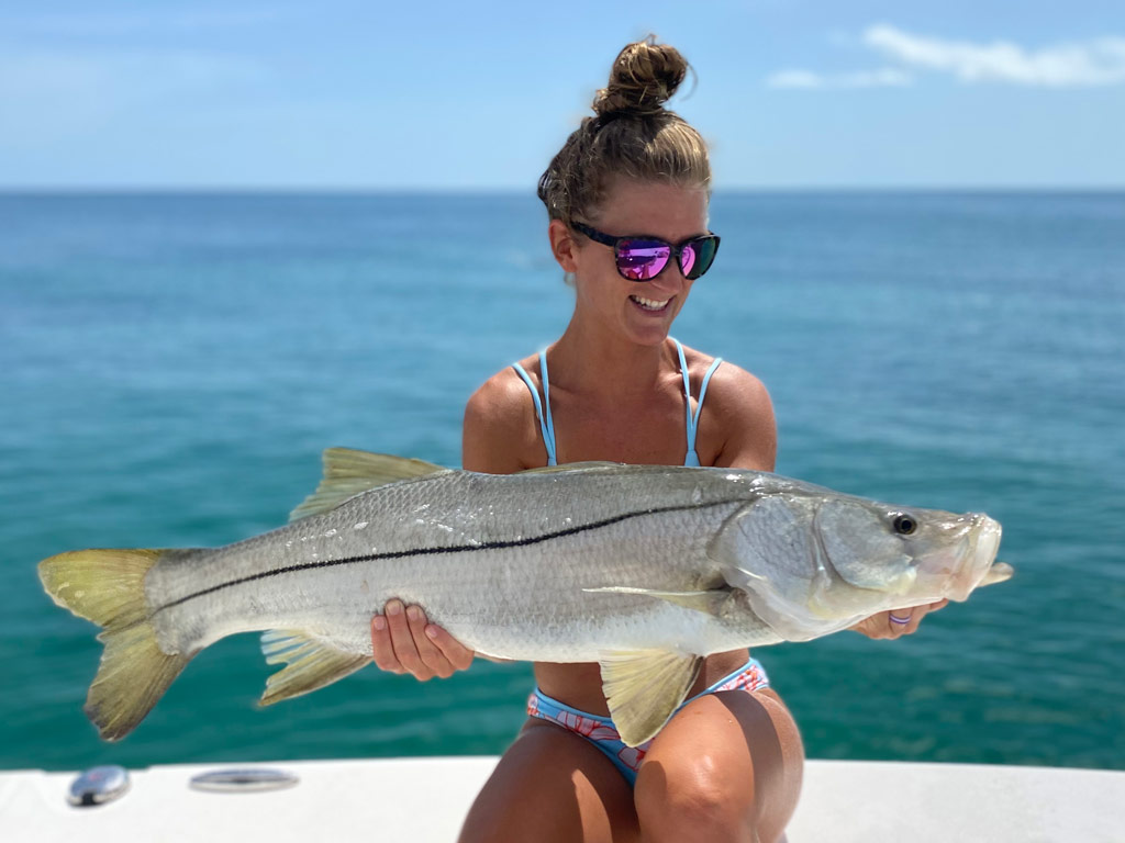 A woman in sunglasses sitting against the side of the boat, smiling and posing for a photo with a huge Snook she caught near Sebastian Inlet, Florida, in the same waters where the state record fish was landed.