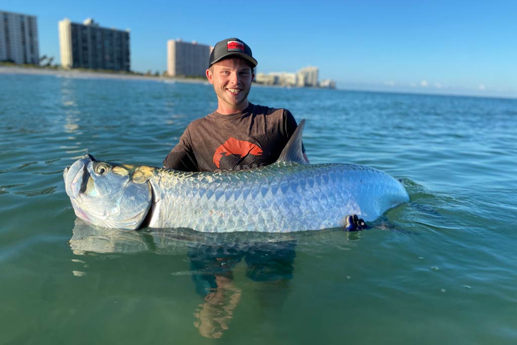 A young smiling angler standing in the water, holding a big Tarpon, with buildings in the far background