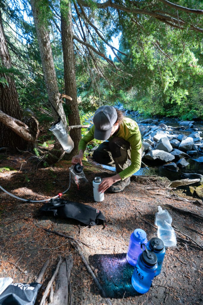 Man filtering water bottles next to a forest stream.