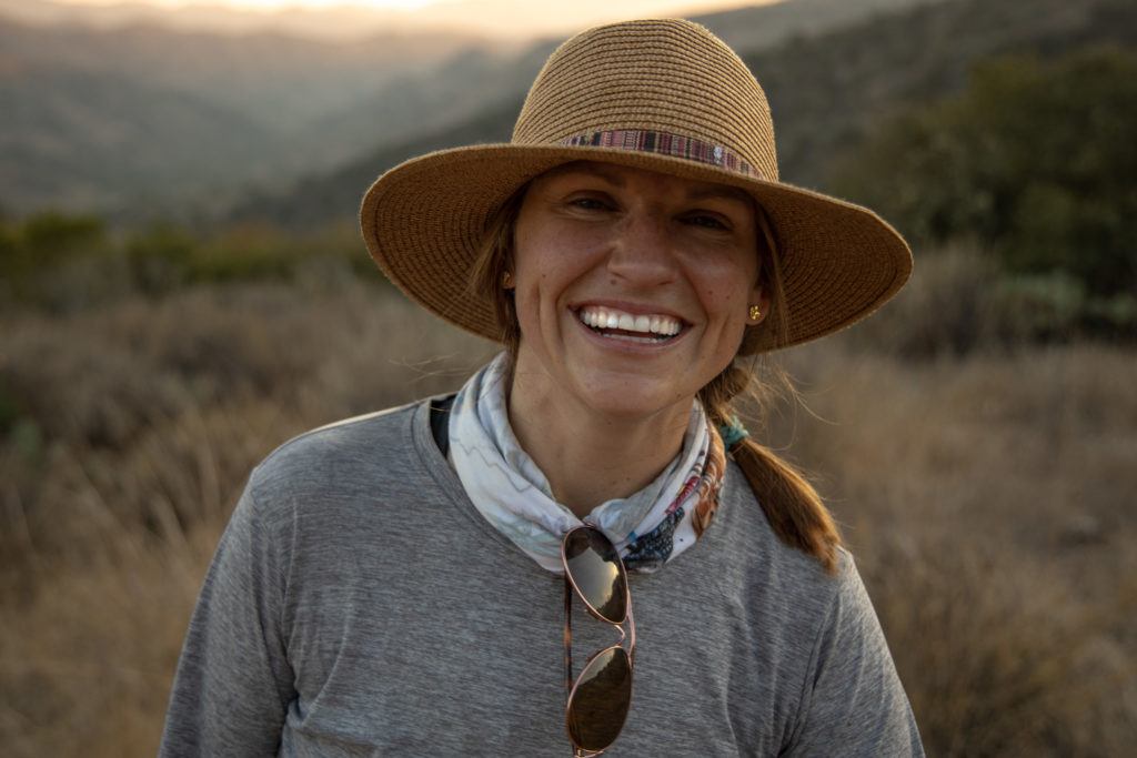 Portait photo of woman wearing sun hat and hiking apparel with setting sun in the background