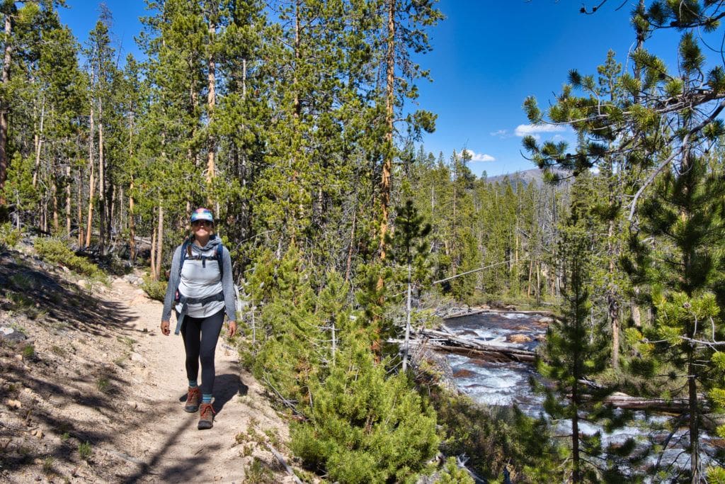 Woman hiking down forested trail next to a river wearing sun protective clothing