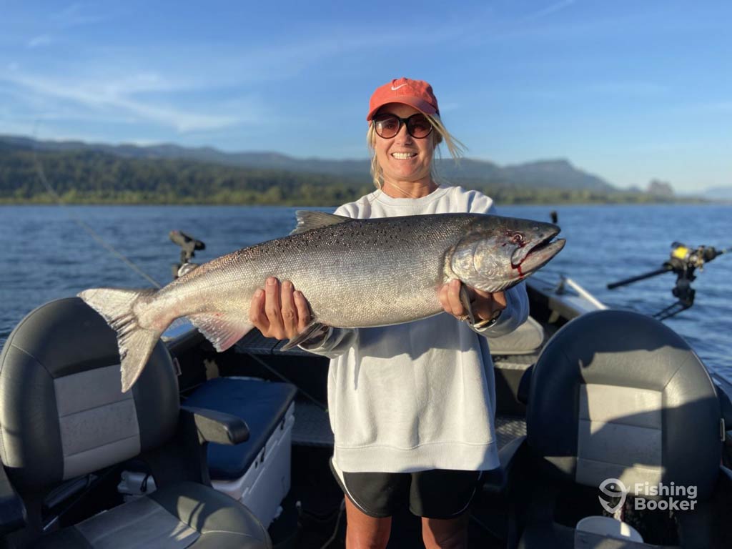 A photo featuring a female angler wearing a cap and a pair of sunglasses while standing on a charter boat and holding a big Salmon caught on a lake 