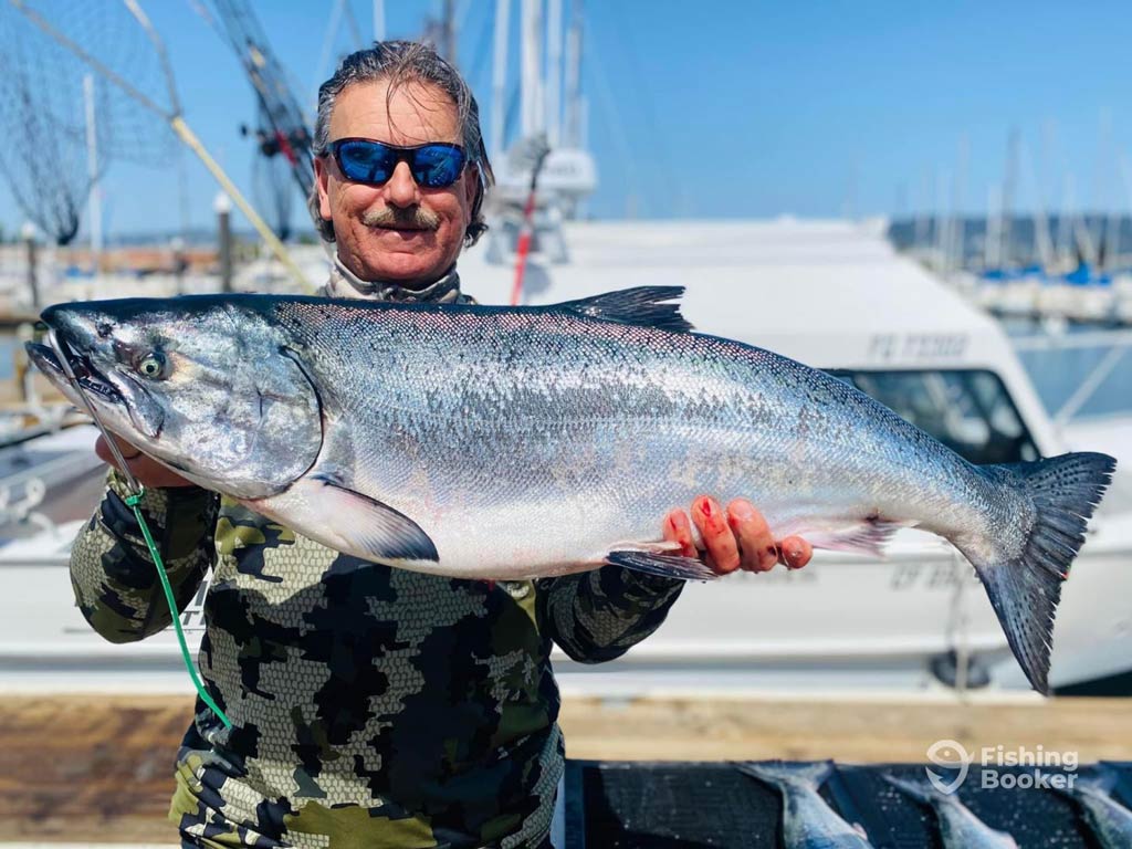 A photo featuring a proud angler wearing a pair of sunglasses with a smile on his face while he is standing on a wooden dock and posing with a big catch in front of charter boats