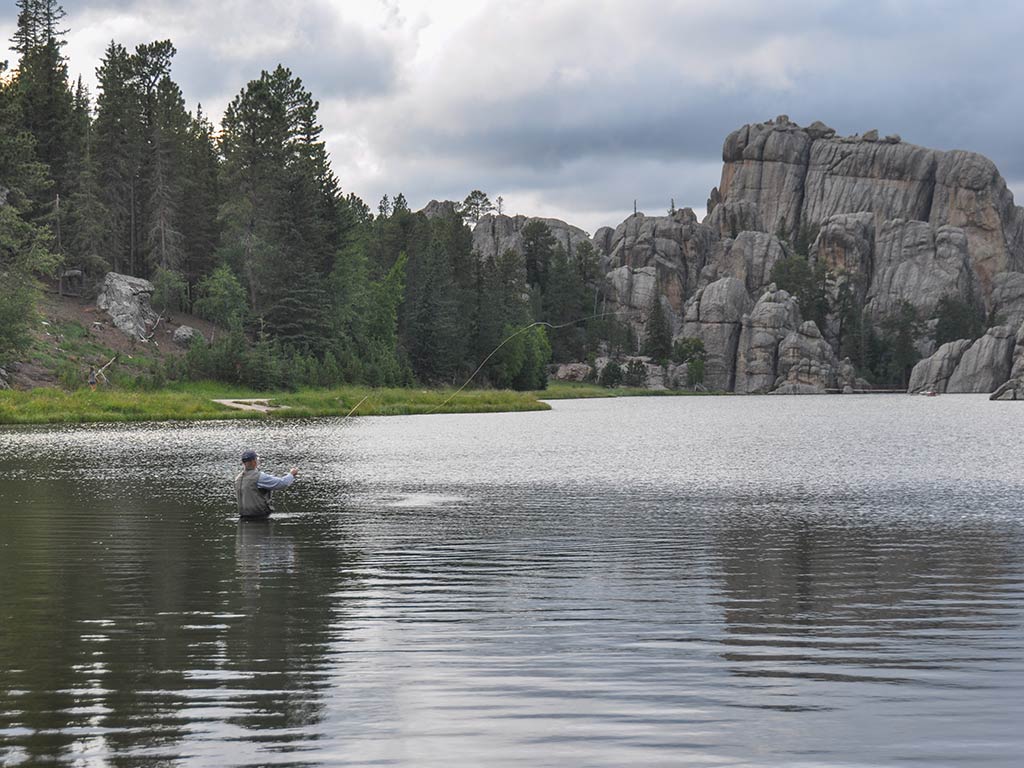 A view across a lake towards a man wade fishing and casting a line into the water in front of some impressive rocks in the distance on a cloudy day