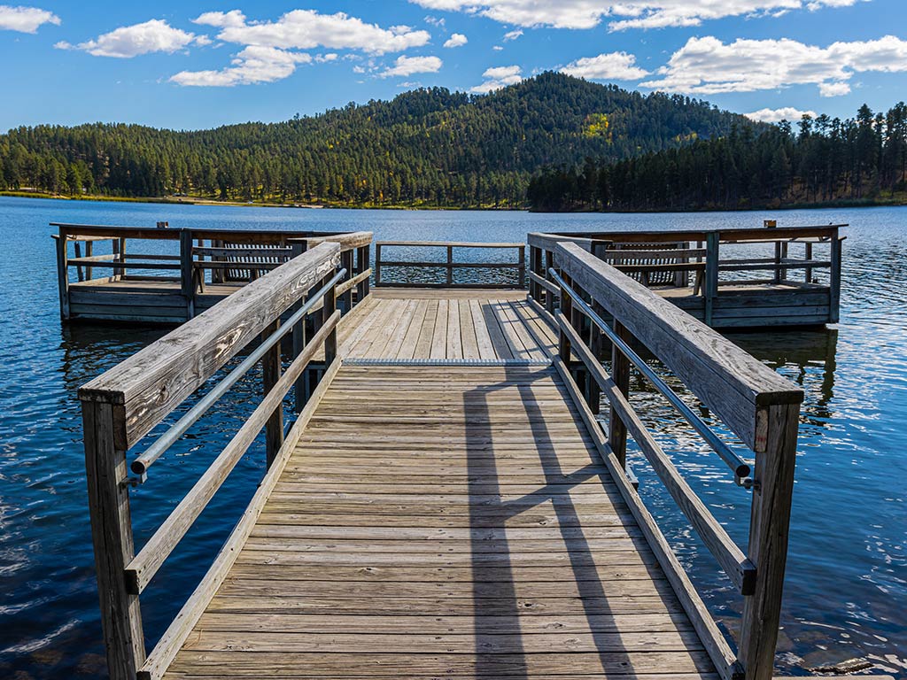 A view towards Stockdale Lake in South Dakota on a clear day across a small, wooden fishing pier, with some hills visible in the distance