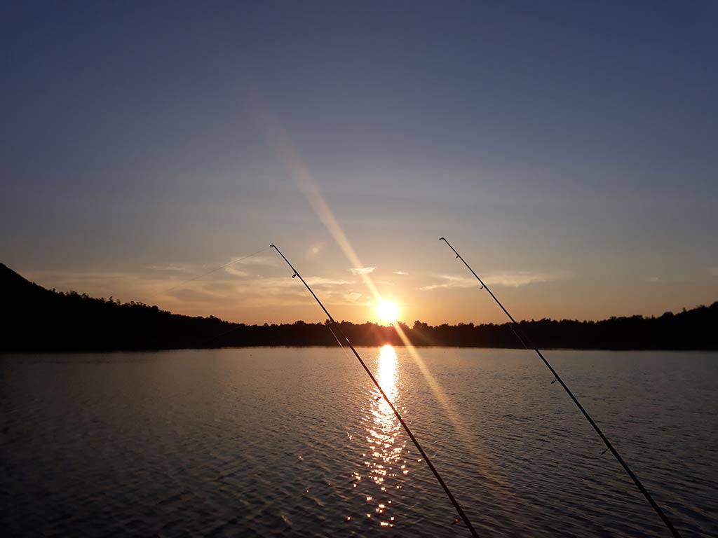 A view towards the sun setting across a lake in South Dakota with the silhouettes of two fishing rods visible in the foreground on a clear day