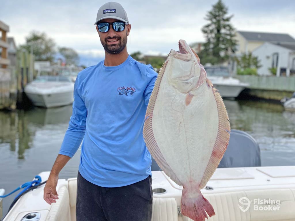 A photo featuring an angler wearing a cap and a pair of sunglasses while standing on a charter boat and posing with a decent-sized Fluke caught on a Memorial Day fishing trip in Montauk