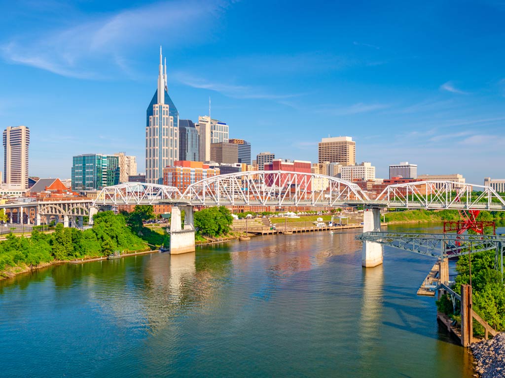 A photo featuring buildings, a bridge, and a river flowing through downtown Nashville on a bright and sunny day