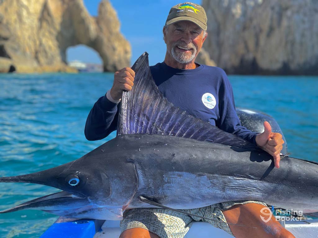 An elderly angler sat on a fishing boat with the famous arch of Cabo San Lucas behind him, with a large Marlin on his lap on a sunny day