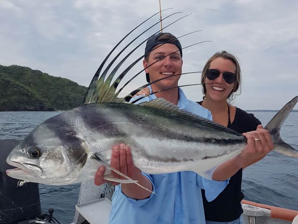 A young man in a backwards baseball cap, holds a Roosterfish up to the camera on a Mexican fishing charter, with a woman standing next to him and some land in the distance on a cloudy day