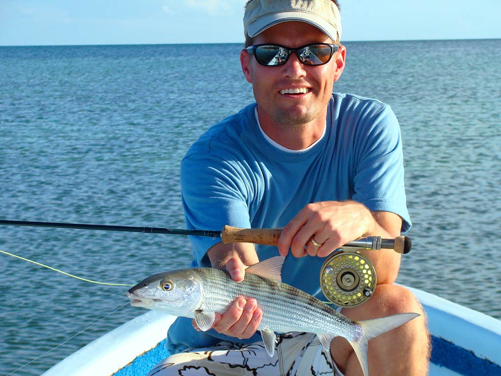 A man in a baseball cap and sunglasses kneeling on a boat in Mexico on a sunny day, holding a Bonefish in one hand and a fly fishing rod in the other