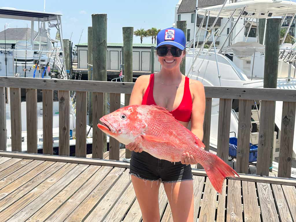 A girl wearing sunglasses and a hat, smiling and standing on a dock while holding a sizeable Red Snapper towards the camera, with boats and dock pillars visible behind her.