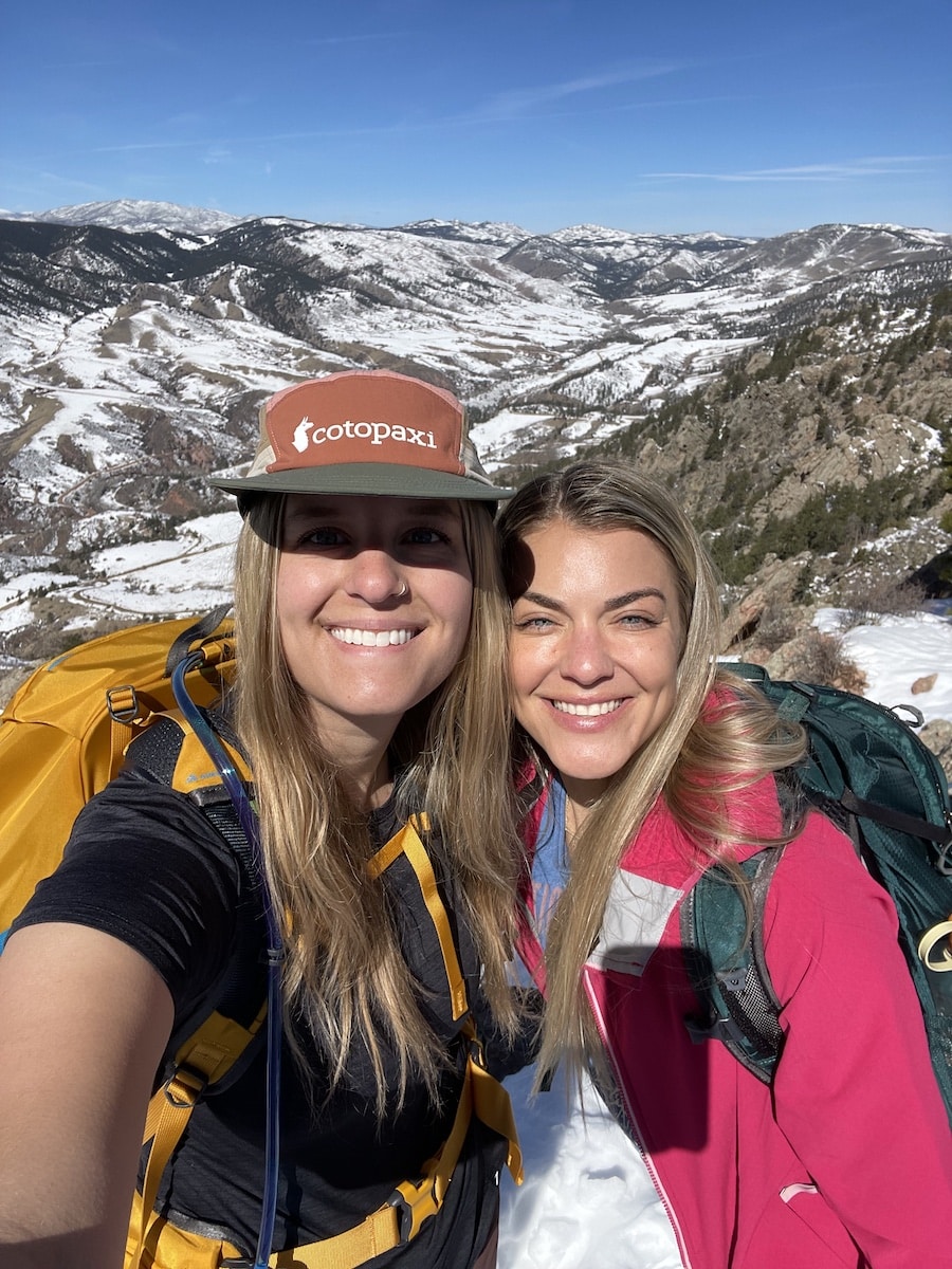 Two women smiling on a hike with snow covered hills in the background