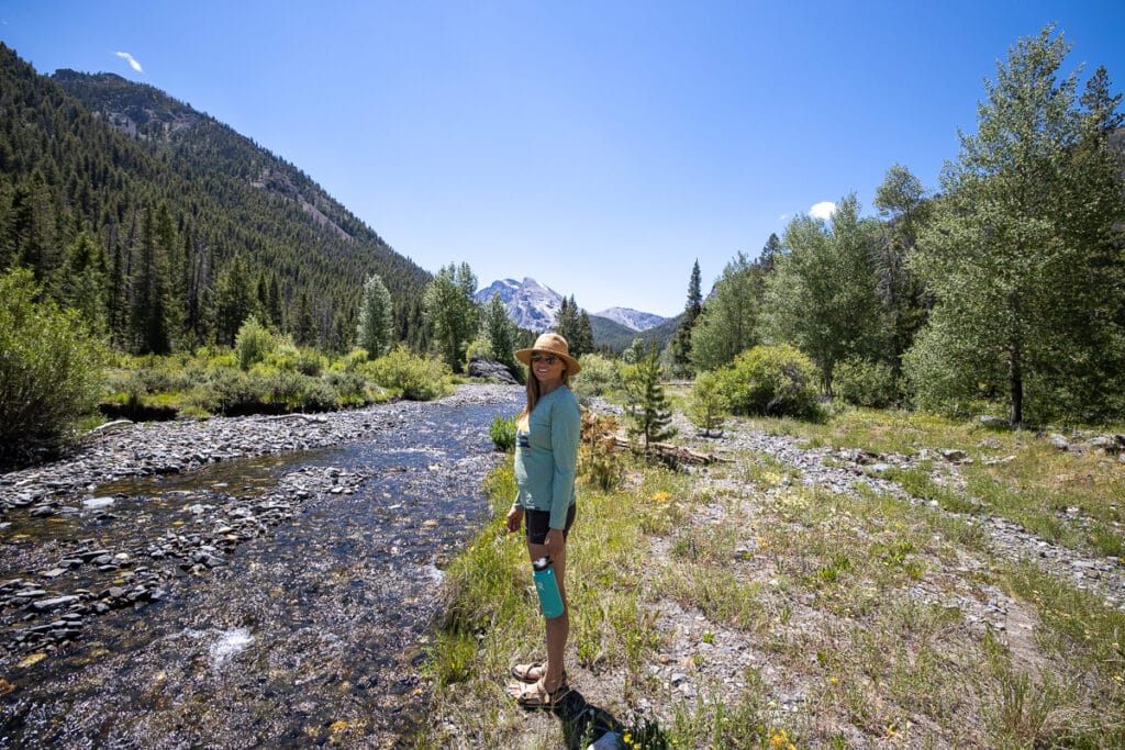 Kristen Bor standing next to a creek surrounding by forested mountains holding a Hydroflask water bottle