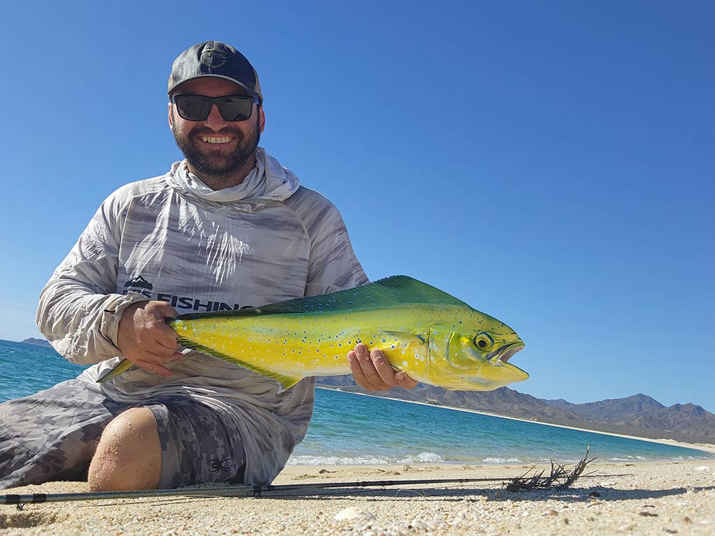 A man in a baseball cap and sunglasses sitting on a beach and holding a mahi Mahi with a fly fishing line visible next to him on a clear day in Mexico