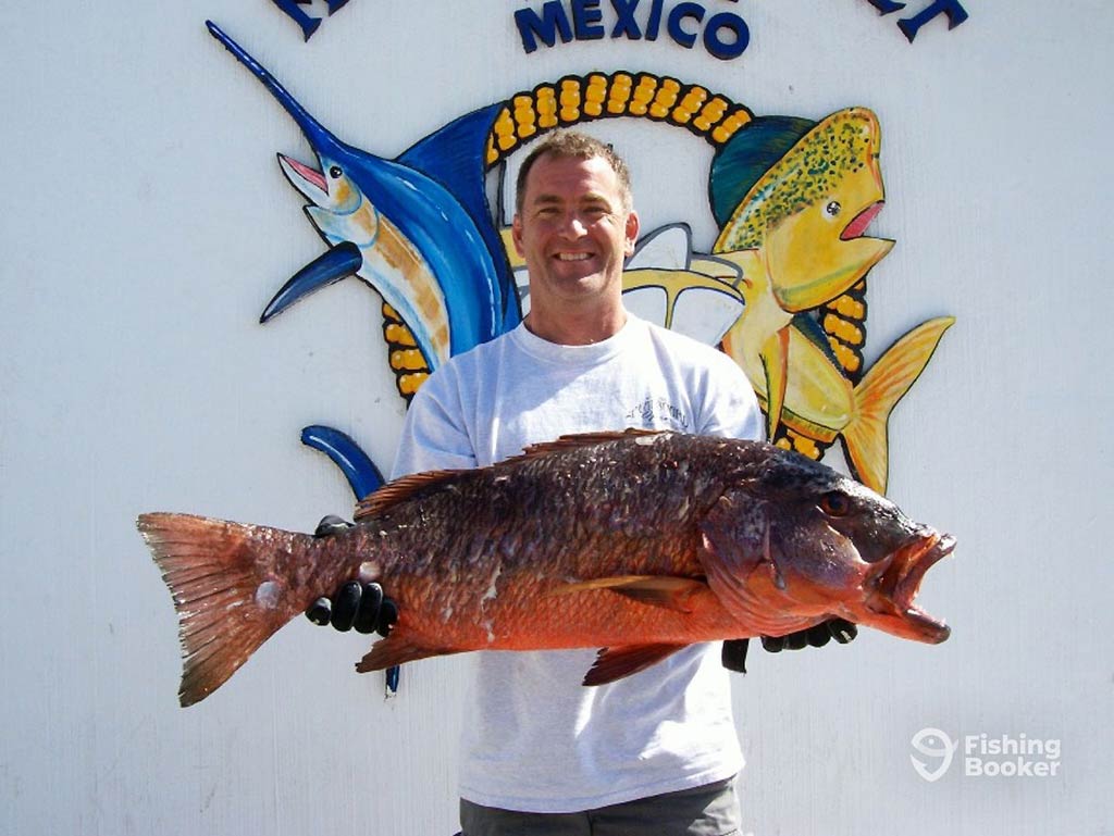 A man smiling and posing in front of a sign after a fishing trip in Mexico, holding a large Grouper