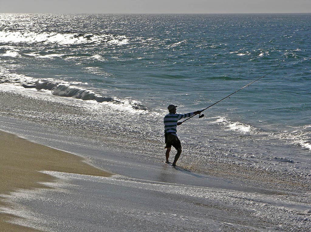 A lone man casts his fishing line into the surf on the Sea of Cortez's coastline at sunset on a bright day