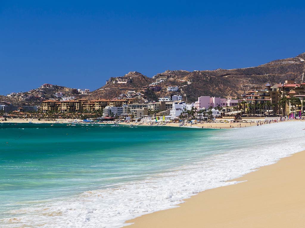 A view along the beach towards the town of Cabo San Lucas with waves crashing in from the turquoise waters on the left of the image on a clear day