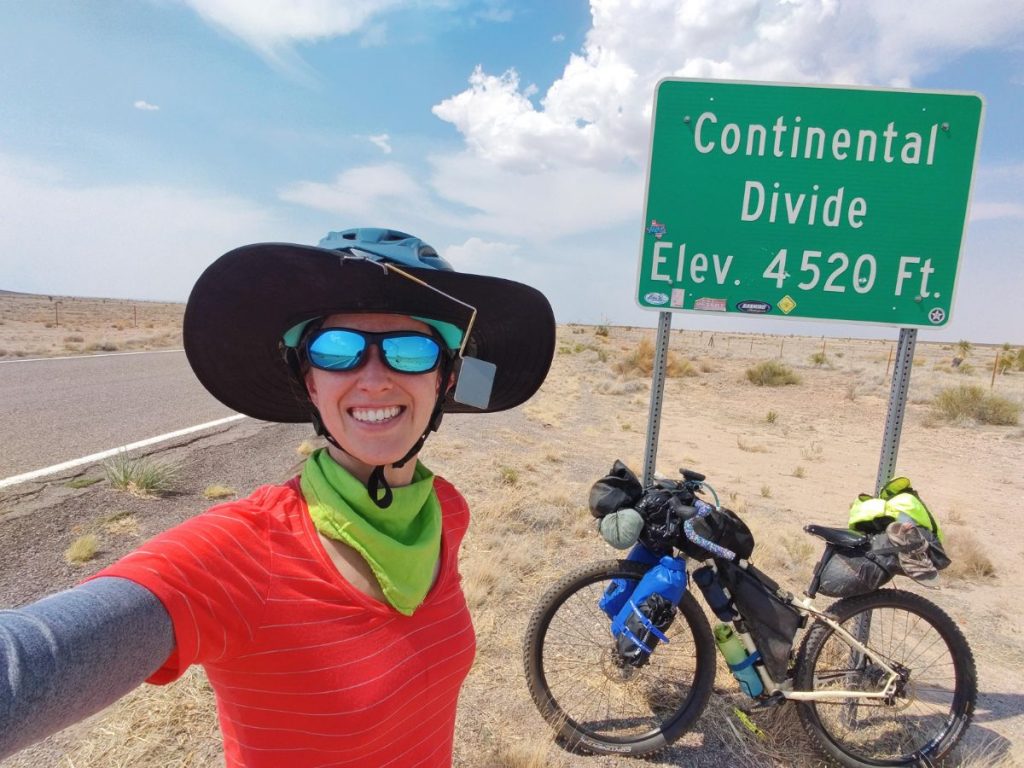 Bikepacker wearing big visor and mirror, with bike leaning against Continental Divide sign in background