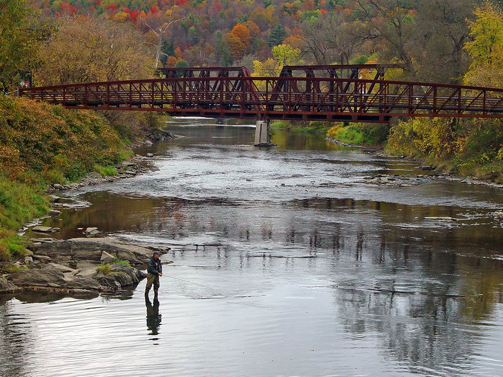 A view from above of a man wade fishing in a river next to a bridge in a river in Vermont in fall