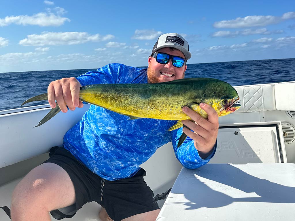 An angler in a baseball cap and sunglasses leaning on a fishing boat and holding a Mahi Mahi on a sunny day