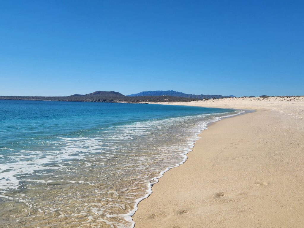A view along a beach on the coast of the Sea of Cortez on a clear day, with calm waters coming into the shore and a few footsteps visible in the foreground