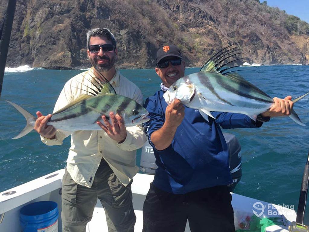 Two men standing on a fishing charter not far from the shore in Costa Rica and holding a colorful Roosterfish each on a clear day