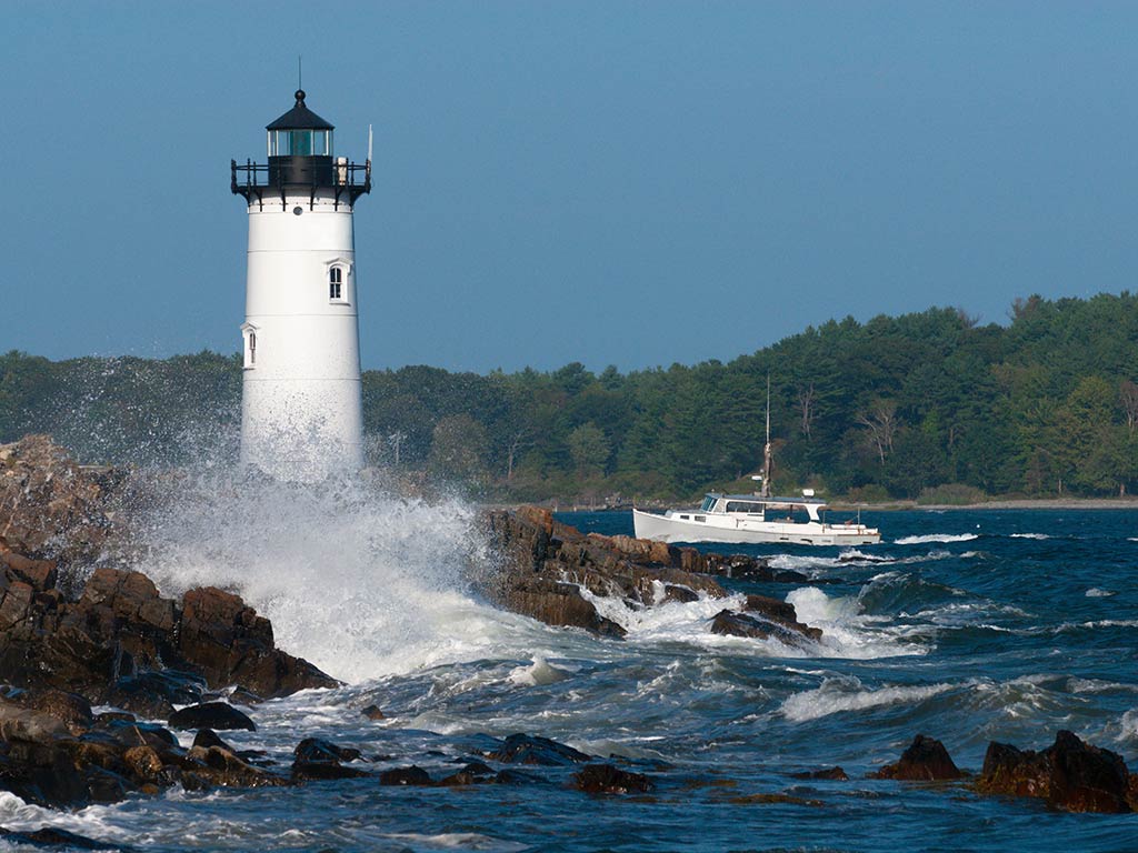 A view across some crashing waves towards a lighthouse in Portsmouth, NH, with a fishing boat visible near it on a clear day