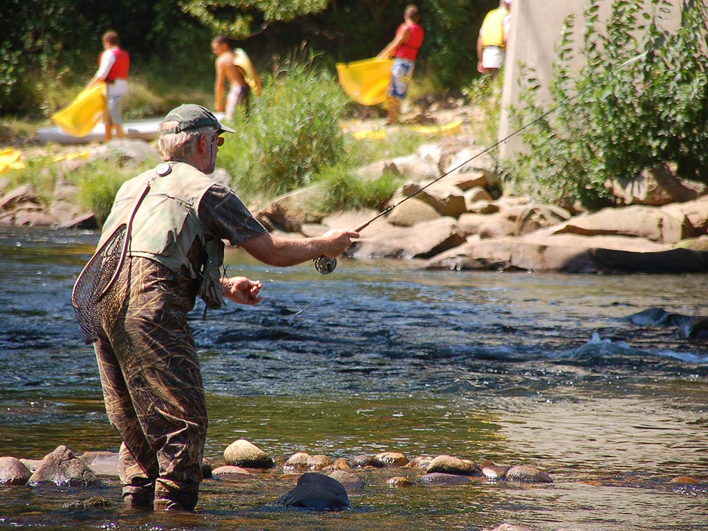 A view from behind of an elderly fly fisherman casting his line into the shallow waters of a stream in New Hampshire on a sunny day