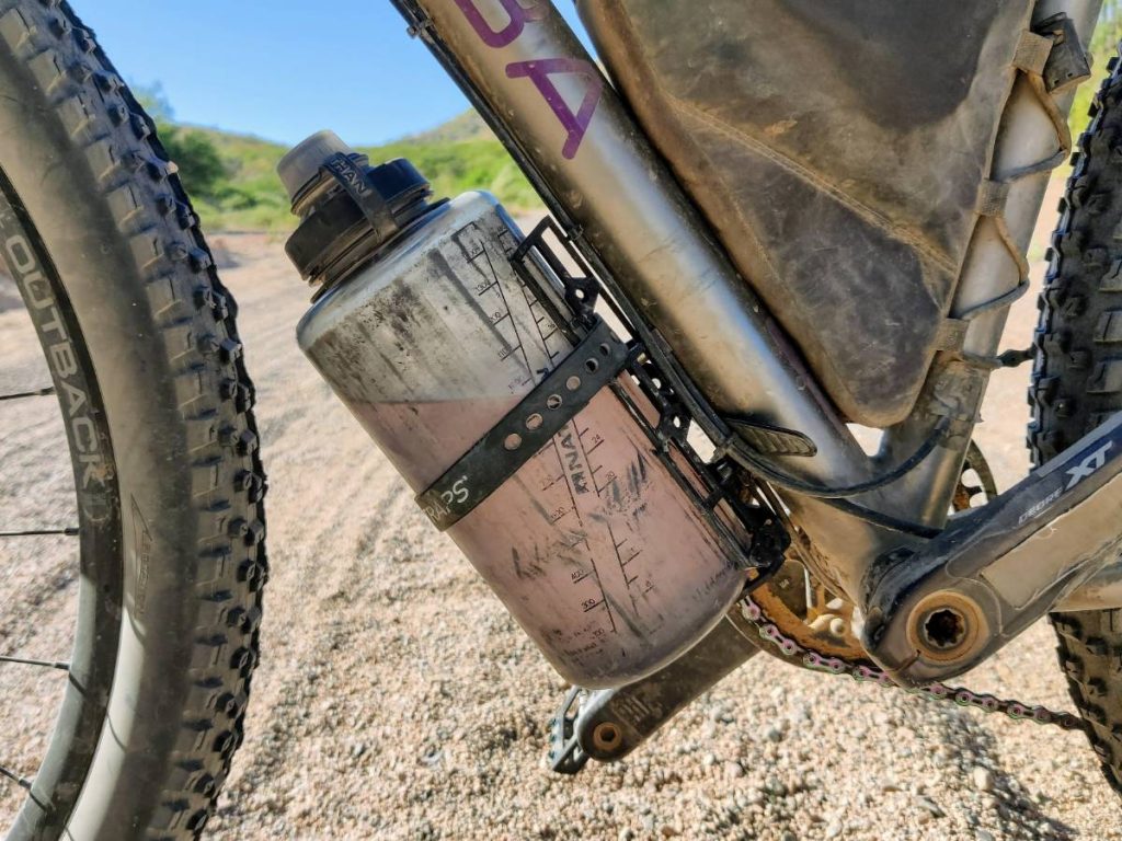 Large plastic bottle in cargo cage on bike downtube against backdrop of sandy road.