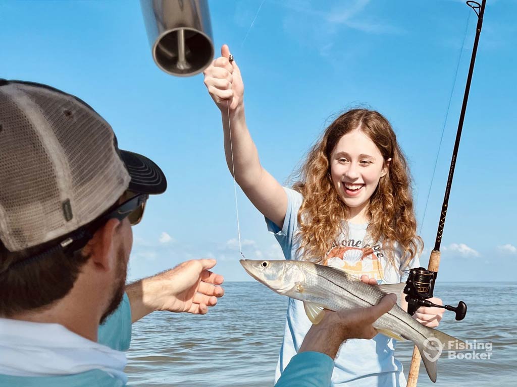 A photo featuring a young female angler happily looking at a fish she caught, with a captain helping her hold the line and the fish on it while they're standing on a charter boat