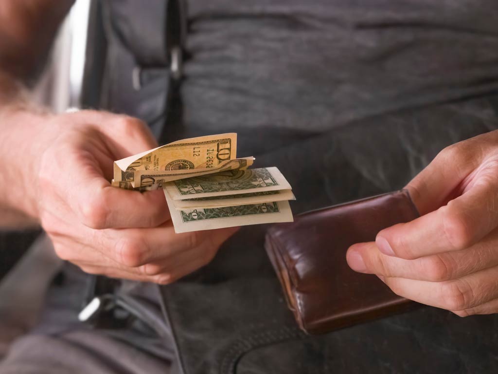 A photo featuring a man while sitting and holding a wallet in one hand and several dollar bills in the other hand thinking about how much to tip a fishing guide
