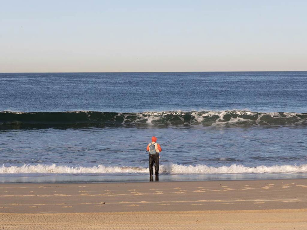 A view from behind of a man surf fishing into the oncoming waves on a beach in Redondo Beach, Florida on a clear day